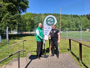 Auf dem Sportgelände des TuS 1920 Oberwinter stehen der 1. Vorsitzende Claus Wiest (rechts) und Thomas Fischer, Verantwortlicher für das Schutz- und Präventionskonzept sowie Ansprechpartner im Verein (links), vor einem Schild mit Vereinslogo und einem Banner zu Gleichberechtigung, Akzeptanz und Vielfalt. Beide tragen Sportkleidung und posieren bei sonnigem Wetter für ein offizielles Vereinsfoto.